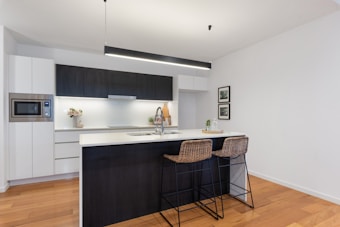 A modern kitchen with a minimalist design featuring white cabinetry and dark wood accents. The central island includes a white countertop and is complemented by two woven bar stools. Appliances like a microwave are integrated into the cabinetry, and decorative elements such as flowers and framed art are present. The lighting is contemporary, using a long, thin pendant light above the island.