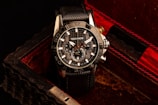 A black and silver wristwatch with a detailed and intricate dial is placed in an ornate, textured box with a dark, luxurious interior featuring red accents. The watch face displays multiple dials and the brand name is visible on it.