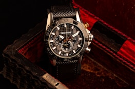 A black and silver wristwatch with a detailed and intricate dial is placed in an ornate, textured box with a dark, luxurious interior featuring red accents. The watch face displays multiple dials and the brand name is visible on it.