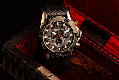 A black and silver wristwatch with a detailed and intricate dial is placed in an ornate, textured box with a dark, luxurious interior featuring red accents. The watch face displays multiple dials and the brand name is visible on it.