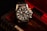 A black and silver wristwatch with a detailed and intricate dial is placed in an ornate, textured box with a dark, luxurious interior featuring red accents. The watch face displays multiple dials and the brand name is visible on it.
