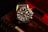 A black and silver wristwatch with a detailed and intricate dial is placed in an ornate, textured box with a dark, luxurious interior featuring red accents. The watch face displays multiple dials and the brand name is visible on it.