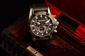 A black and silver wristwatch with a detailed and intricate dial is placed in an ornate, textured box with a dark, luxurious interior featuring red accents. The watch face displays multiple dials and the brand name is visible on it.