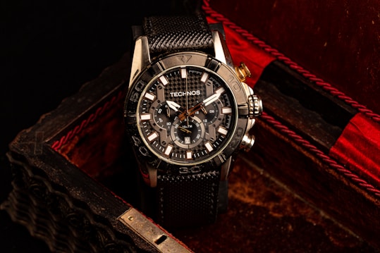 A black and silver wristwatch with a detailed and intricate dial is placed in an ornate, textured box with a dark, luxurious interior featuring red accents. The watch face displays multiple dials and the brand name is visible on it.