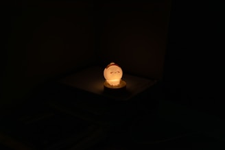 A small glowing night lamp with a cute sleeping face design sits on a flat surface. The lamp emits a warm yellow-orange light, casting a soft glow around it, while the rest of the surroundings remain dark and shadowy.
