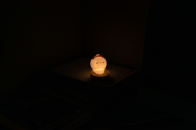 A small glowing night lamp with a cute sleeping face design sits on a flat surface. The lamp emits a warm yellow-orange light, casting a soft glow around it, while the rest of the surroundings remain dark and shadowy.