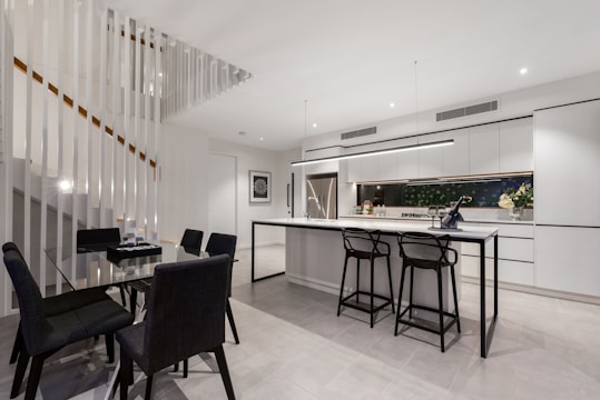 A modern kitchen and dining area with sleek design elements. The kitchen features white cabinetry with black accents, a large stainless steel refrigerator, and a long island counter with black bar stools. The dining area has a glass table with black chairs. A staircase with vertical white slats is visible on the left.