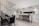 A modern kitchen and dining area with sleek design elements. The kitchen features white cabinetry with black accents, a large stainless steel refrigerator, and a long island counter with black bar stools. The dining area has a glass table with black chairs. A staircase with vertical white slats is visible on the left.