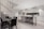 A modern kitchen and dining area with sleek design elements. The kitchen features white cabinetry with black accents, a large stainless steel refrigerator, and a long island counter with black bar stools. The dining area has a glass table with black chairs. A staircase with vertical white slats is visible on the left.