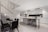 A modern kitchen and dining area with sleek design elements. The kitchen features white cabinetry with black accents, a large stainless steel refrigerator, and a long island counter with black bar stools. The dining area has a glass table with black chairs. A staircase with vertical white slats is visible on the left.