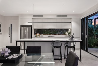A modern kitchen with a sleek design featuring a large island with bar stools, stainless steel appliances, and a glass door opening to a garden. The countertops are white, and the cabinets are minimalistic. The space is well-lit with recessed lighting and a decorative light fixture above the island.
