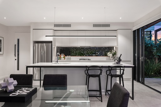 A modern kitchen with a sleek design featuring a large island with bar stools, stainless steel appliances, and a glass door opening to a garden. The countertops are white, and the cabinets are minimalistic. The space is well-lit with recessed lighting and a decorative light fixture above the island.