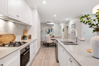 A modern kitchen with light-colored cabinetry and a gray countertop. The kitchen features a gas stove and built-in oven on the left side, and a sink with a modern faucet on the right. A vase with green foliage adds a touch of nature. The backsplash has a herringbone tile pattern, and there are decorative wooden cutting boards. In the background, a dining area with white chairs and a circular rug is visible.