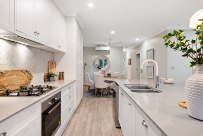 A modern kitchen with light-colored cabinetry and a gray countertop. The kitchen features a gas stove and built-in oven on the left side, and a sink with a modern faucet on the right. A vase with green foliage adds a touch of nature. The backsplash has a herringbone tile pattern, and there are decorative wooden cutting boards. In the background, a dining area with white chairs and a circular rug is visible.