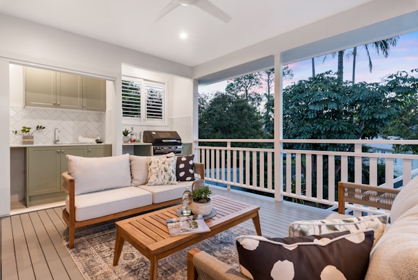 A well-furnished outdoor patio with a cozy seating area consisting of a sofa and chairs, all with decorative cushions. A coffee table holds a small potted plant and magazines. In the background, an outdoor kitchen area with cabinetry and a barbecue grill is visible. The area is surrounded by lush greenery and large trees, offering a serene view.