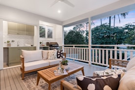 A well-furnished outdoor patio with a cozy seating area consisting of a sofa and chairs, all with decorative cushions. A coffee table holds a small potted plant and magazines. In the background, an outdoor kitchen area with cabinetry and a barbecue grill is visible. The area is surrounded by lush greenery and large trees, offering a serene view.