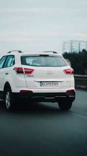 A white Hyundai SUV is driving on a road. The vehicle's rear view is visible, and it has a license plate with the number KL 69 H 6969. The sky appears overcast, and there is some motion blur, suggesting the car is in motion.