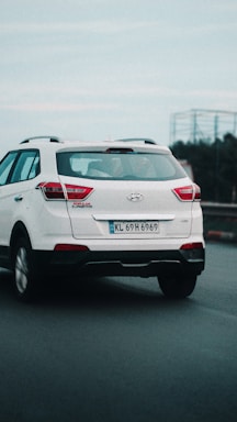A white Hyundai SUV is driving on a road. The vehicle's rear view is visible, and it has a license plate with the number KL 69 H 6969. The sky appears overcast, and there is some motion blur, suggesting the car is in motion.