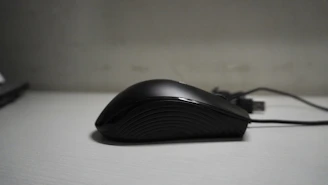 A sleek, black computer mouse with a smooth, ergonomic design is positioned on a light gray surface. The mouse has a subtle texture on its side for better grip, and an attached USB cable extends from the back.