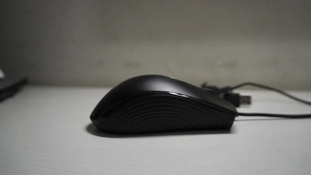 A sleek, black computer mouse with a smooth, ergonomic design is positioned on a light gray surface. The mouse has a subtle texture on its side for better grip, and an attached USB cable extends from the back.