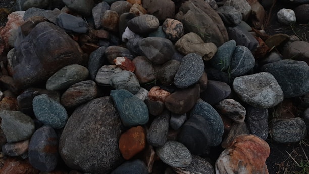A collection of variously sized and colored stones is piled together. The stones exhibit a range of earthy tones, including grays, browns, blues, and reds. They appear smooth and weathered, suggesting exposure to natural elements over time.