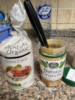A packet of organic corn cakes sits on a kitchen counter next to an open jar of natural crunchy peanut butter with a black-handled knife inserted into it. In the background, there's a large bottle of water and part of another kitchen item on the left.