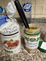 A packet of organic corn cakes sits on a kitchen counter next to an open jar of natural crunchy peanut butter with a black-handled knife inserted into it. In the background, there's a large bottle of water and part of another kitchen item on the left.