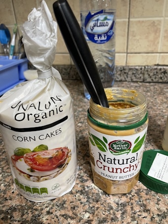 A packet of organic corn cakes sits on a kitchen counter next to an open jar of natural crunchy peanut butter with a black-handled knife inserted into it. In the background, there's a large bottle of water and part of another kitchen item on the left.