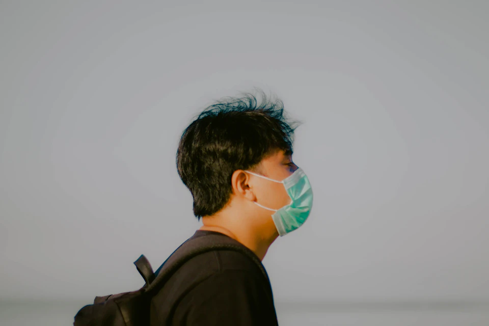 Person wearing a face mask stands with backpack on left side, gazing off to right