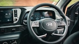 A car's interior focusing on the steering wheel and dashboard. The dashboard features a digital display with gauges indicating speed and fuel. The infotainment screen is visible, showing options for radio and media settings. The steering wheel has control buttons and the manufacturer's logo.