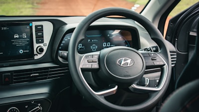A car's interior focusing on the steering wheel and dashboard. The dashboard features a digital display with gauges indicating speed and fuel. The infotainment screen is visible, showing options for radio and media settings. The steering wheel has control buttons and the manufacturer's logo.