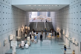 A modern museum interior features smooth concrete walls with quadratic perforations and a reflective glass floor. Ancient artifacts and sculptures are displayed on both sides. Visitors walk and observe the exhibits, with a staircase leading to an upper level in the background.