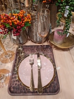 An elegant dining setup with a light pink plate edged in gold, placed on a textured brown mat. A gold knife and fork are neatly arranged on the plate. Surrounding the setting are vibrant orange, red, and green berries in decorative metal vases, alongside a tall champagne flute. A pink candle sits in a gold dish nearby, and the overall arrangement is placed on a light wooden table.