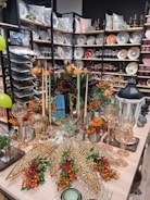 An elegantly arranged display featuring decorative plates, glassware, and autumn-themed floral arrangements. Shelves are filled with a variety of plates, bowls, and pillows, creating a luxurious home decor atmosphere. In the foreground, an assortment of gold ornamental leaves and glass candle holders are complemented by vibrant orange and red flowers.