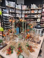 An elegantly arranged display featuring decorative plates, glassware, and autumn-themed floral arrangements. Shelves are filled with a variety of plates, bowls, and pillows, creating a luxurious home decor atmosphere. In the foreground, an assortment of gold ornamental leaves and glass candle holders are complemented by vibrant orange and red flowers.