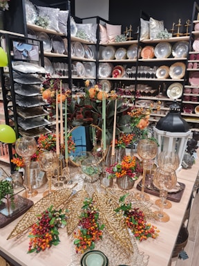 An elegantly arranged display featuring decorative plates, glassware, and autumn-themed floral arrangements. Shelves are filled with a variety of plates, bowls, and pillows, creating a luxurious home decor atmosphere. In the foreground, an assortment of gold ornamental leaves and glass candle holders are complemented by vibrant orange and red flowers.