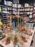 An elegantly arranged display featuring decorative plates, glassware, and autumn-themed floral arrangements. Shelves are filled with a variety of plates, bowls, and pillows, creating a luxurious home decor atmosphere. In the foreground, an assortment of gold ornamental leaves and glass candle holders are complemented by vibrant orange and red flowers.