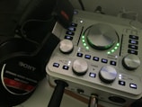 A close-up of an audio interface with various knobs, buttons, and indicators on its panel. The interface is accompanied by a pair of Sony dynamic stereo headphones, positioned to the left. The interface has LED lights illuminating in green around the central knob.