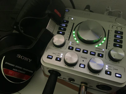 A close-up of an audio interface with various knobs, buttons, and indicators on its panel. The interface is accompanied by a pair of Sony dynamic stereo headphones, positioned to the left. The interface has LED lights illuminating in green around the central knob.