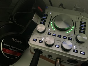 A close-up of an audio interface with various knobs, buttons, and indicators on its panel. The interface is accompanied by a pair of Sony dynamic stereo headphones, positioned to the left. The interface has LED lights illuminating in green around the central knob.