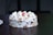 A round cake topped with white cream and garnished with almond slices, featuring a single red cherry in the center. The cake rests on a dark wooden table and conveys a sense of simplicity and elegance.