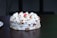 A round cake topped with white cream and garnished with almond slices, featuring a single red cherry in the center. The cake rests on a dark wooden table and conveys a sense of simplicity and elegance.