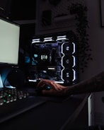 A dark, modern computer setup featuring a gaming PC with RGB lighting and a visible GeForce RTX graphics card. A hand is using a mouse, and a keyboard is partially visible. Plants are seen in the background on a wall shelf, adding a touch of greenery to the room.