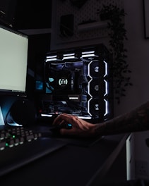 A dark, modern computer setup featuring a gaming PC with RGB lighting and a visible GeForce RTX graphics card. A hand is using a mouse, and a keyboard is partially visible. Plants are seen in the background on a wall shelf, adding a touch of greenery to the room.