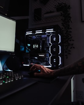 A dark, modern computer setup featuring a gaming PC with RGB lighting and a visible GeForce RTX graphics card. A hand is using a mouse, and a keyboard is partially visible. Plants are seen in the background on a wall shelf, adding a touch of greenery to the room.