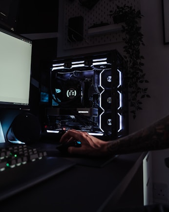 A dark, modern computer setup featuring a gaming PC with RGB lighting and a visible GeForce RTX graphics card. A hand is using a mouse, and a keyboard is partially visible. Plants are seen in the background on a wall shelf, adding a touch of greenery to the room.