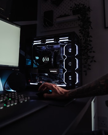 A dark, modern computer setup featuring a gaming PC with RGB lighting and a visible GeForce RTX graphics card. A hand is using a mouse, and a keyboard is partially visible. Plants are seen in the background on a wall shelf, adding a touch of greenery to the room.