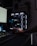 A dark, modern computer setup featuring a gaming PC with RGB lighting and a visible GeForce RTX graphics card. A hand is using a mouse, and a keyboard is partially visible. Plants are seen in the background on a wall shelf, adding a touch of greenery to the room.