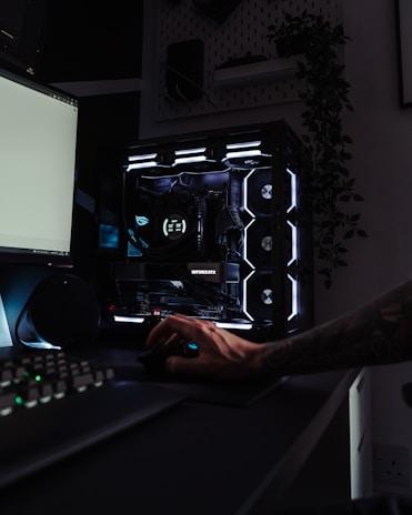 A dark, modern computer setup featuring a gaming PC with RGB lighting and a visible GeForce RTX graphics card. A hand is using a mouse, and a keyboard is partially visible. Plants are seen in the background on a wall shelf, adding a touch of greenery to the room.