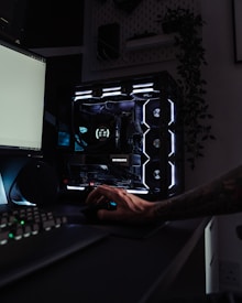 A dark, modern computer setup featuring a gaming PC with RGB lighting and a visible GeForce RTX graphics card. A hand is using a mouse, and a keyboard is partially visible. Plants are seen in the background on a wall shelf, adding a touch of greenery to the room.